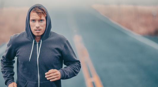 Mann in dunklem Hoodie joggt auf einer nebligen Straße. Sein Gesichtsausdruck zeigt Entschlossenheit, während er fokussiert nach vorne blickt.