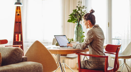 Frau in Business-Kleidung sitzt an einem runden Holztisch und arbeitet am Laptop. Der Raum ist stilvoll eingerichtet mit modernen Möbeln und Pflanzen, Tageslicht strömt durch große Fenster.