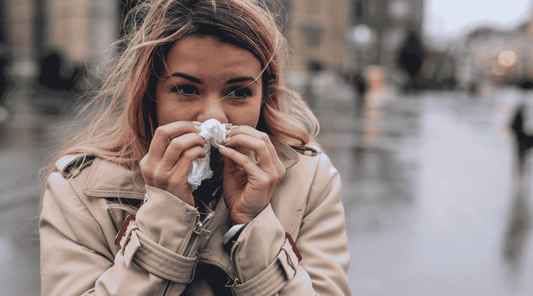 Junge Frau steht im Freien und schnäuzt sich mit einem Taschentuch die Nase, umgeben von einem trüben, regnerischen Stadtbild – Symbolbild für Erkältung bei kaltem Wetter.