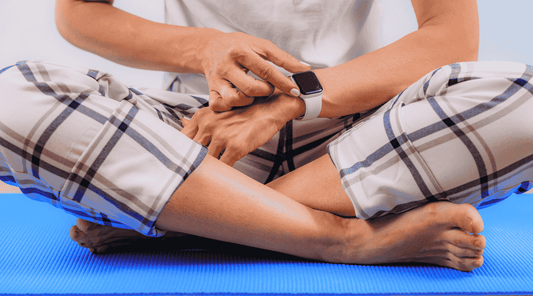 Eine Person sitzt im Schneidersitz auf einer blauen Yogamatte und trägt eine karierte Pyjamahose. Sie berührt ihre Smartwatch am Handgelenk
